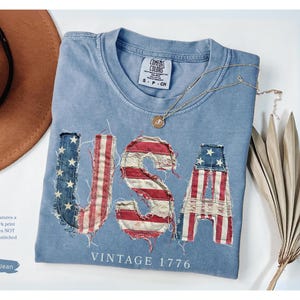 May include: A blue jean colored t-shirt with the word "USA" in a faux patchwork print design. The letters are made to look like the American flag. The shirt also has the words "VINTAGE 1776" printed on it. A brown hat and gold necklace are also in the image.