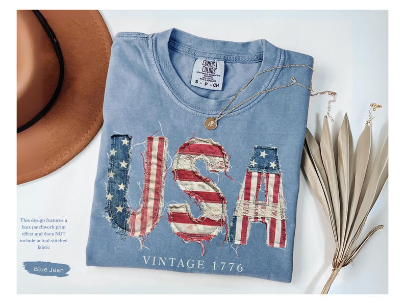 Puede incluir: Una camiseta azul vaquera con la palabra "USA" en un dise&ntilde;o de estampado de patchwork falso. Las letras est&aacute;n hechas para parecerse a la bandera estadounidense. La camisa tambi&eacute;n tiene las palabras "VINTAGE 1776" impresas. Un sombrero marr&oacute;n y un collar dorado tambi&eacute;n est&aacute;n en la imagen.