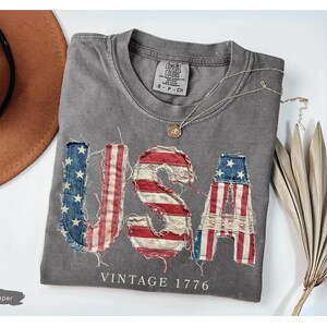 Puede incluir: Una camiseta gris con la palabra "USA" en un dise&ntilde;o desgastado de la bandera estadounidense. Tambi&eacute;n aparece el texto "VINTAGE 1776". Un sombrero marr&oacute;n y un collar plateado tambi&eacute;n est&aacute;n en la imagen.