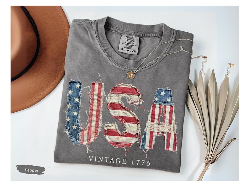 Puede incluir: Una camiseta gris con la palabra "USA" en un dise&ntilde;o desgastado de la bandera estadounidense. Tambi&eacute;n aparece el texto "VINTAGE 1776". Un sombrero marr&oacute;n y un collar plateado tambi&eacute;n est&aacute;n en la imagen.