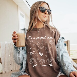 May include: A brown t-shirt with white text that reads "It's a perfect day for a miracle." The shirt features a variety of small white illustrations including a heart, a dove, a stethoscope, footprints, a rainbow, and a hand with a flower.