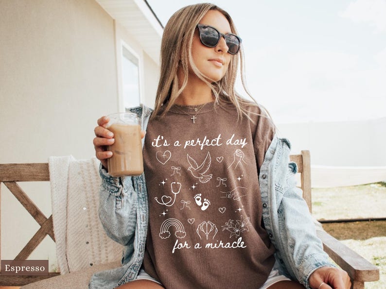 May include: A brown t-shirt with white text that reads "It's a perfect day for a miracle." The shirt features a variety of small white illustrations including a heart, a dove, a stethoscope, footprints, a rainbow, and a hand with a flower.