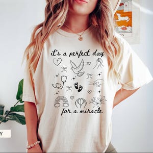 May include: A white t-shirt with the text "it's a perfect day for a miracle" printed in black. The text is surrounded by small black illustrations of hearts, a dove, a stethoscope, footprints, a rainbow, and flowers.