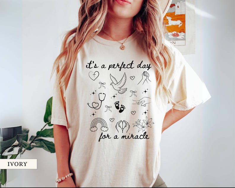 May include: A white t-shirt with the text "it's a perfect day for a miracle" printed in black. The text is surrounded by small black illustrations of hearts, a dove, a stethoscope, footprints, a rainbow, and flowers.