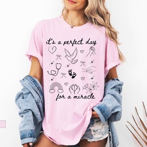 May include: Pink t-shirt with the text "it's a perfect day for a miracle" and various small black and white illustrations including a dove, a rainbow, a stethoscope, a heart, and footprints.