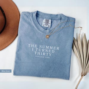 May include: Blue jean colored t-shirt with the text "THE SUMMER I TURNED THIRTY VINTAGE 1996" in white. A gold necklace with a pendant rests on the shirt. A brown hat and dried plant are also in the image.