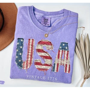 Puede incluir: Una camiseta violeta Comfort Colors con la palabra "USA" en un dise&ntilde;o de bandera americana desgastada. El texto "VINTAGE 1776" est&aacute; debajo de las letras. Un collar dorado y un sombrero marr&oacute;n tambi&eacute;n est&aacute;n en la imagen.