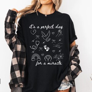 May include: Black t-shirt with white text that reads "it's a perfect day for a miracle". The shirt features a variety of white illustrations including a dove, a stethoscope, footprints, a rainbow, and flowers.