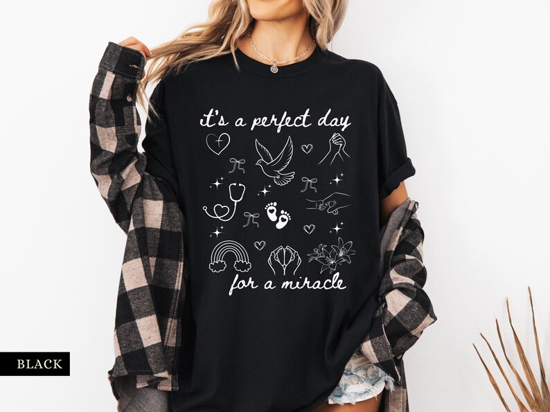 May include: Black t-shirt with white text that reads "it's a perfect day for a miracle". The shirt features a variety of white illustrations including a dove, a stethoscope, footprints, a rainbow, and flowers.