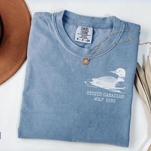 May include: A blue jean colored t-shirt with the text "STUPID CANADIAN WOLF BIRD" and a graphic of a loon. A gold necklace with a pendant and a brown hat are also in the image. The shirt is a Comfort Colors brand.