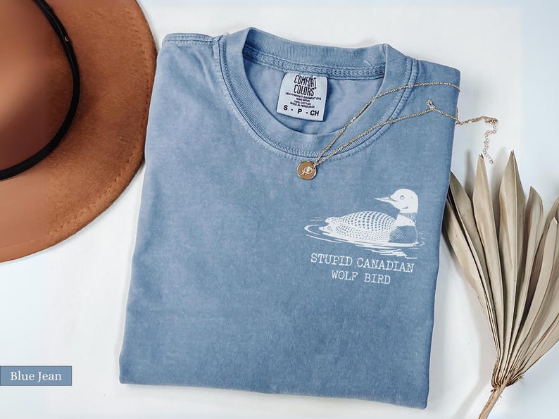 May include: A blue jean colored t-shirt with the text "STUPID CANADIAN WOLF BIRD" and a graphic of a loon. A gold necklace with a pendant and a brown hat are also in the image. The shirt is a Comfort Colors brand.