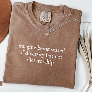 May include: Espresso-colored t-shirt with the text "imagine being scared of diversity but not dictatorship." A silver necklace with a small pendant rests on the shirt. The shirt is folded on a white surface.