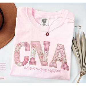 May include: A light pink t-shirt with the letters "CNA" in a patchwork design. The shirt also has the words "certified nursing assistant" in script. A brown hat and necklace are next to the shirt.