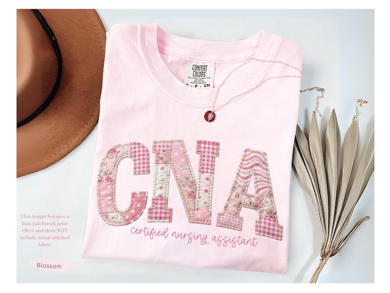 May include: A pale pink t-shirt featuring the letters "CNA" in a patchwork design. The shirt also has the words "certified nursing assistant" in script. A brown hat and necklace are next to the shirt.