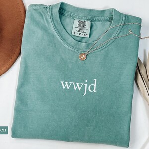 Puede incluir: Camiseta verde claro Comfort Colors con el texto "wwjd" en blanco. Un collar de oro con un colgante est&aacute; sobre la camiseta. Un sombrero marr&oacute;n y una planta seca est&aacute;n en el fondo.