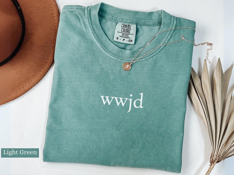 Puede incluir: Camiseta verde claro Comfort Colors con el texto "wwjd" en blanco. Un collar de oro con un colgante est&aacute; sobre la camiseta. Un sombrero marr&oacute;n y una planta seca est&aacute;n en el fondo.