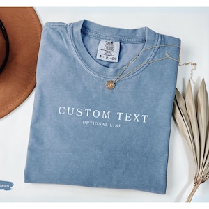 May include: A blue jean colored t-shirt with the words "CUSTOM TEXT OPTIONAL LINE" printed in white. A gold necklace and a brown hat are next to the shirt. A dried plant is on the right side of the shirt.