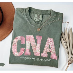 May include: A moss green t-shirt featuring the letters "CNA" in floral and gingham patchwork. The words "certified nursing assistant" are below. A brown hat and a necklace are also visible in the image.