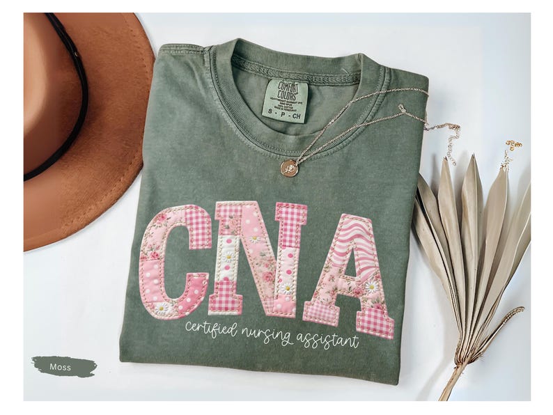 May include: A moss green t-shirt featuring the letters "CNA" in floral and gingham patchwork. The words "certified nursing assistant" are below. A brown hat and a necklace are also visible in the image.