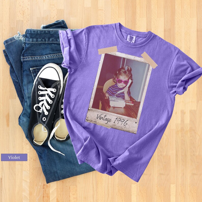 Pu&ograve; includere: Una maglietta viola con una stampa fotografica vintage, abbinata a jeans blu e scarpe da ginnastica di tela nere. La foto &egrave; attaccata alla maglietta e mostra un bambino con occhiali da sole che usa un telefono. Il testo "Vintage 1996" &egrave; stampato sulla foto.