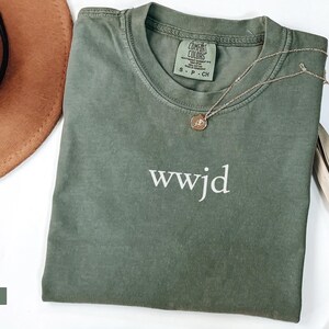 Puede incluir: Una camiseta verde musgo con el texto blanco "wwjd". Un collar dorado con un colgante est&aacute; sobre la camiseta. Un sombrero marr&oacute;n est&aacute; en la esquina superior izquierda. La camiseta es de la marca "Comfort Colors".