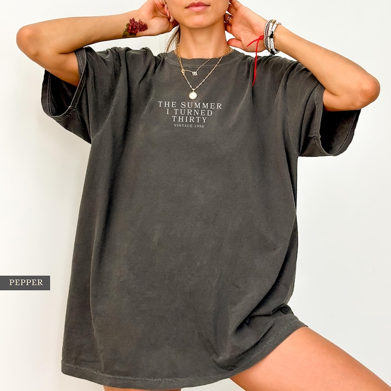 May include: Dark gray oversized t-shirt with the text "THE SUMMER I TURNED THIRTY VINTAGE 1996" in white. The shirt has short sleeves and a relaxed fit. The shirt is made of a soft, comfortable material.