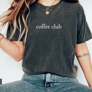 May include: Dark grey t-shirt with the words "coffee club" in white. The shirt is worn with blue jeans. The model is wearing a necklace and bracelets. The shirt is a casual style.
