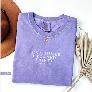 May include: A violet Comfort Colors t-shirt with the text "THE SUMMER I TURNED THIRTY VINTAGE 1996" in white. A gold necklace with a pendant rests on the shirt. A brown hat and dried plant are in the background.