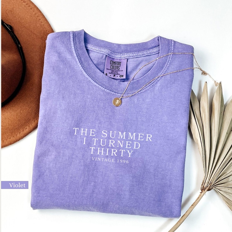 May include: A violet Comfort Colors t-shirt with the text "THE SUMMER I TURNED THIRTY VINTAGE 1996" in white. A gold necklace with a pendant rests on the shirt. A brown hat and dried plant are in the background.