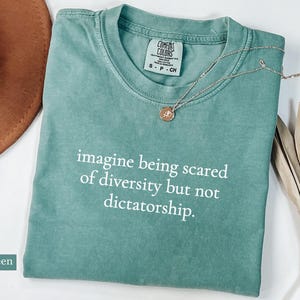 May include: Light green t-shirt with the text "imagine being scared of diversity but not dictatorship." A gold necklace with a pendant rests on the shirt. A brown hat is visible in the background.