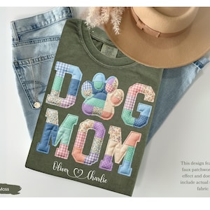 May include: Moss green t-shirt with the words "DOG MOM" in a patchwork design, featuring a paw print. The shirt includes the names "Oliver & Charlie" and is paired with blue jeans and a tan hat.