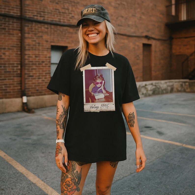 Pu&ograve; includere: T-shirt nero con stampa fotografica vintage Polaroid. La foto mostra un bambino con occhiali da sole e la scritta "Vintage 1996". La persona indossa un berretto nero.