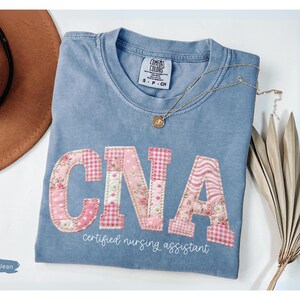 May include: A blue jean coloured t-shirt with the letters "CNA" appliqu&eacute;d in pink floral and gingham patterns. The words "certified nursing assistant" are printed below the letters in a script font. A gold necklace and a brown hat are also in the image.