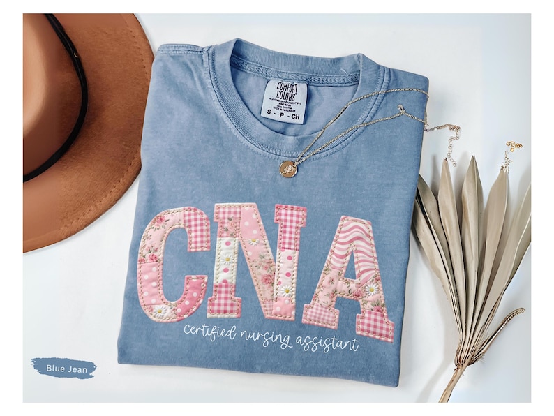 May include: A blue jean coloured t-shirt with the letters "CNA" appliqu&eacute;d in pink floral and gingham patterns. The words "certified nursing assistant" are printed below the letters in a script font. A gold necklace and a brown hat are also in the image.