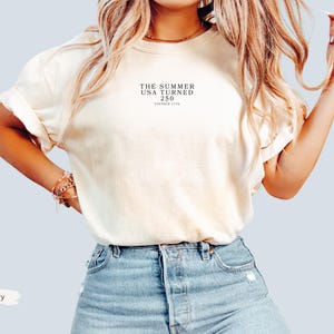 May include: Ivory-colored t-shirt with rolled sleeves and the text "THE SUMMER USA TURNED 250 VINTAGE 1776" printed in black. The shirt is paired with blue denim shorts. The word "Ivory" is written in a white rectangle.