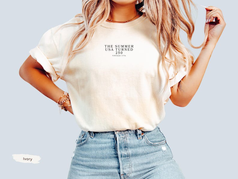 May include: Ivory-colored t-shirt with rolled sleeves and the text "THE SUMMER USA TURNED 250 VINTAGE 1776" printed in black. The shirt is paired with blue denim shorts. The word "Ivory" is written in a white rectangle.