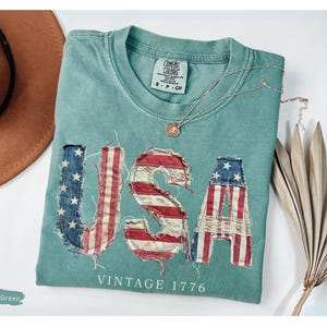 Puede incluir: Camiseta verde claro con la palabra "USA" en un dise&ntilde;o desgastado de la bandera estadounidense. El texto "VINTAGE 1776" est&aacute; debajo de la palabra "USA". Un sombrero marr&oacute;n y un collar dorado tambi&eacute;n est&aacute;n en la imagen.