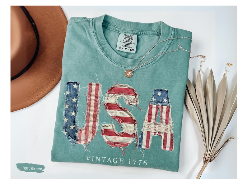 Puede incluir: Camiseta verde claro con la palabra "USA" en un dise&ntilde;o desgastado de la bandera estadounidense. El texto "VINTAGE 1776" est&aacute; debajo de la palabra "USA". Un sombrero marr&oacute;n y un collar dorado tambi&eacute;n est&aacute;n en la imagen.