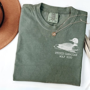 May include: Moss green t-shirt with a graphic of a loon and the text "STUPID CANADIAN WOLF BIRD". A gold necklace and a brown hat are also visible. The shirt is made of a soft material.