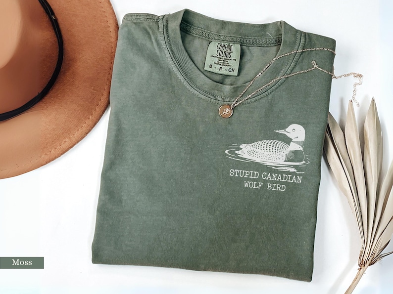 May include: Moss green t-shirt with a graphic of a loon and the text "STUPID CANADIAN WOLF BIRD". A gold necklace and a brown hat are also visible. The shirt is made of a soft material.