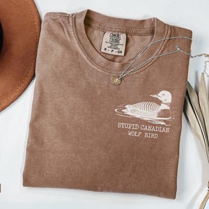 May include: Espresso-colored t-shirt with a white graphic of a loon and the text "STUPID CANADIAN WOLF BIRD." A silver necklace with a small pendant rests on the shirt. A brown hat and dried plant are also in the image.