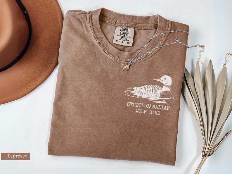 May include: Espresso-colored t-shirt with a white graphic of a loon and the text "STUPID CANADIAN WOLF BIRD." A silver necklace with a small pendant rests on the shirt. A brown hat and dried plant are also in the image.