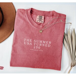 May include: Crimson-colored t-shirt with the text "THE SUMMER USA TURNED 250 VINTAGE 1776" in white. A brown hat and a silver necklace with a pendant are next to the shirt. Dried decorative plants are on the right.