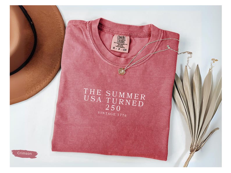 May include: Crimson-colored t-shirt with the text "THE SUMMER USA TURNED 250 VINTAGE 1776" in white. A brown hat and a silver necklace with a pendant are next to the shirt. Dried decorative plants are on the right.