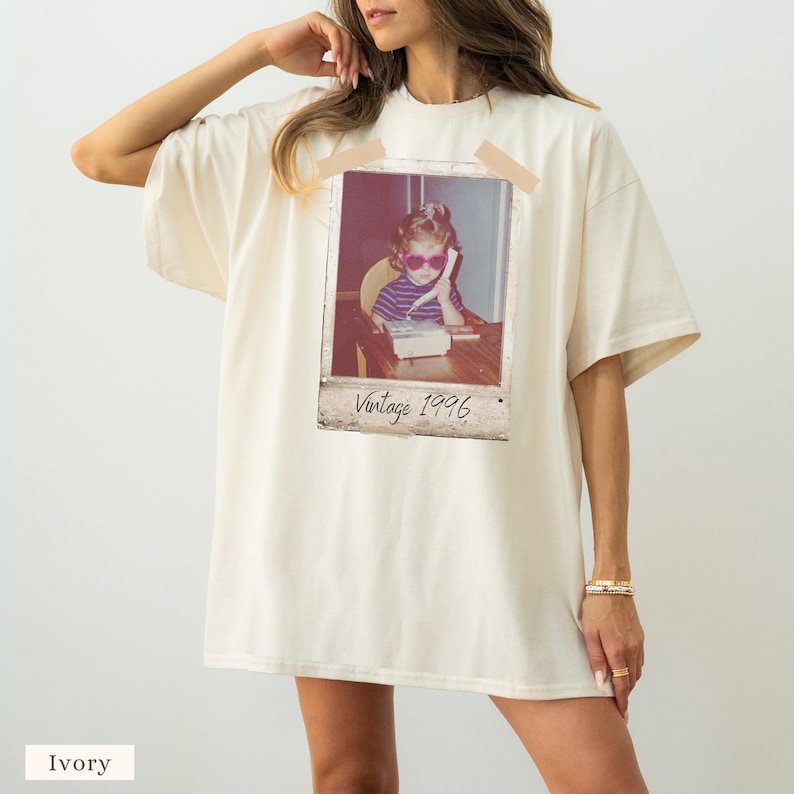 Pu&ograve; includere: T-shirt oversize color avorio con stampa fotografica Polaroid vintage. La stampa mostra un bambino con occhiali da sole che usa un telefono, con la scritta "Vintage 1996". La maglietta ha maniche corte e una vestibilit&agrave; ampia.