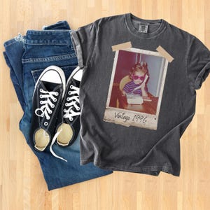 Pu&ograve; includere: Un outfit casual con una maglietta grigio antracite con stampa fotografica vintage, jeans blu e scarpe da ginnastica nere in tela. La maglietta presenta una foto di un bambino con occhiali da sole e un telefono, con la scritta "Vintage 1996".