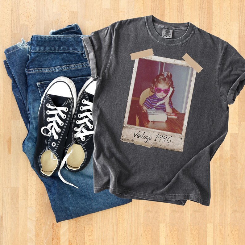 Pu&ograve; includere: Un outfit casual con una maglietta grigio antracite con stampa fotografica vintage, jeans blu e scarpe da ginnastica nere in tela. La maglietta presenta una foto di un bambino con occhiali da sole e un telefono, con la scritta "Vintage 1996".