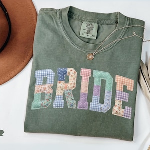 May include: A moss green t-shirt with the word "BRIDE" in patchwork letters. The letters are made of various floral and gingham fabrics. A gold necklace with a pendant is on the shirt. A brown hat and dried plant are also in the image.