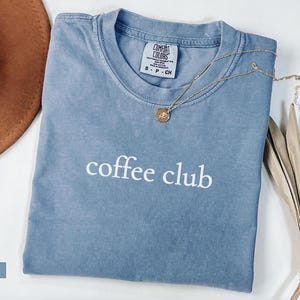 May include: A blue jean coloured t-shirt with the words "coffee club" in white. A gold necklace with a pendant rests on the shirt. A brown hat and dried palm leaves are also in the image.