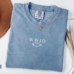 May include: A blue jean colored t-shirt with the text "WWJD" and a fish symbol in white. A gold necklace with a pendant rests on the shirt. A brown hat and dried plant are in the background.